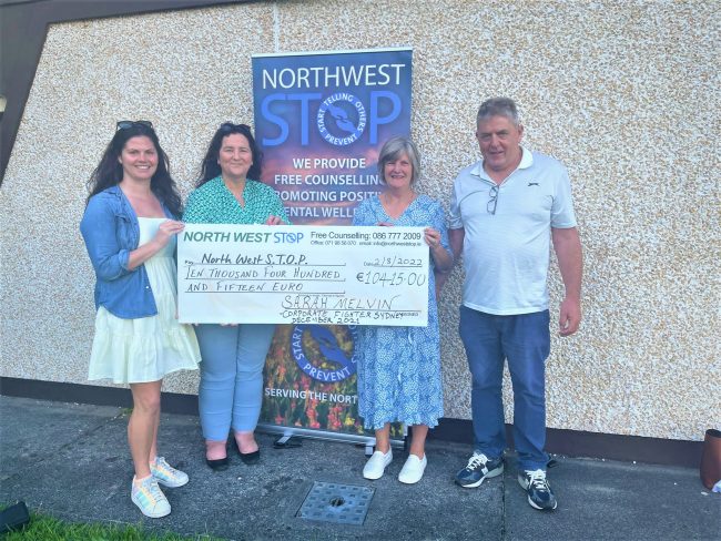 Four people stand in front of a pebble-dashed wall. Three of them hold a large novelty presentation cheque. Behind them is a pop-up banner which reads North West STOP: we provide free counselling...